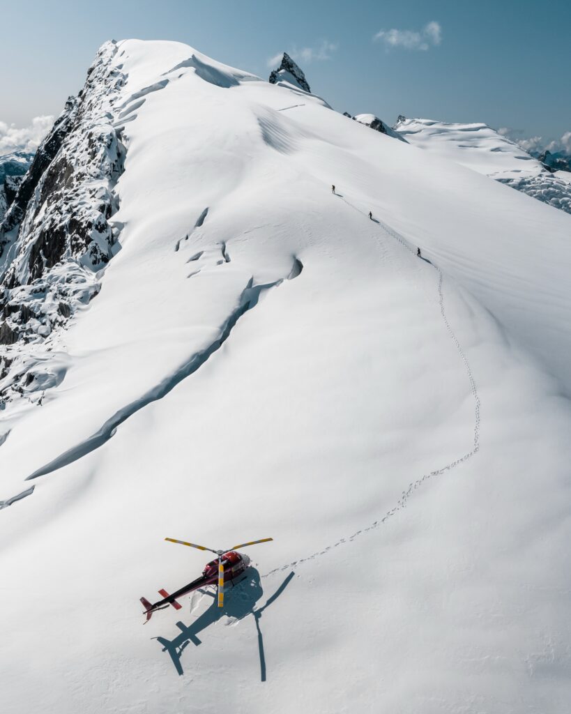 Red helicopter below climbers ascending a snow-covered alpine ridge