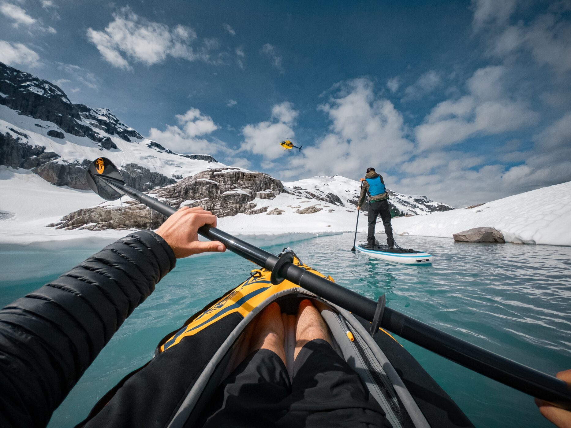 Kayaker following a paddleboarder across a turquoise alpine lake as a helicopter flies overhead